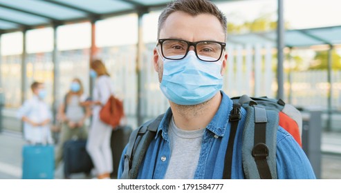 Portrait of handsome Caucasian man in glasses and medical mask looking behind, then at camera and standing at bus stop or train station. Wife with kids and suitcases on blurred background. Pandemic.