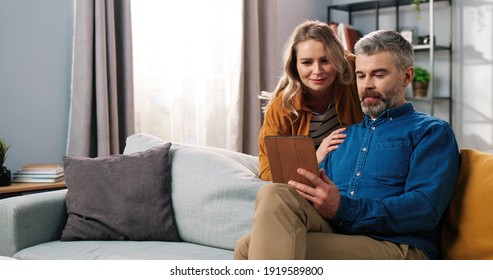 Portrait of handsome Caucasian middle-aged bearded grey-haired man sitting on sofa in room surfing internet on tablet browsing and choosing something with his beautiful happy wife, family concept