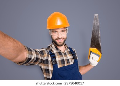 Portrait of handsome cheerful master in overall, shirt, hardhat, uniform shooting selfie on front camera, showing saw in arm, having leisure fun, isolated on grey background