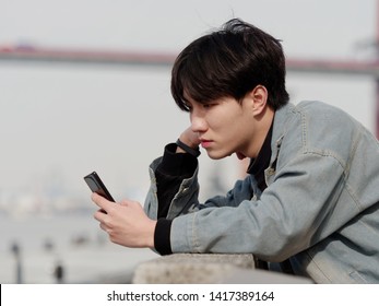 Portrait Of A Handsome Chinese Young Man Holding His Mobile Phone, Looking For Something Interesting In Internet, Mobile Phone And The Internet Take Up A Lot Of Teenager's Time.