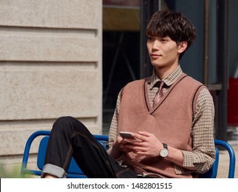 Portrait Of A Handsome Chinese Young Man With Korean Style Clothes Sitting And Looking Away With Mobile Phone In Hand, Male Fashion, Cool Asian Young Man Lifestyle.