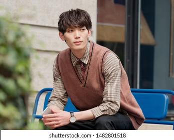 Portrait Of A Handsome Chinese Young Man With Korean Style Clothes Sitting And Looking Away With Mobile Phone In Hand, Male Fashion, Cool Asian Young Man Lifestyle.