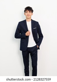 Portrait Of Handsome Chinese Young Man In Dark Blue Leisure Suit Posing Against White Wall Background. Hand In Pocket With Another Hand On Suit And Smiling At Camera, Looks Smart And Confident.