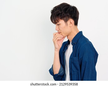 Portrait Of Handsome Chinese Young Man With Curly Black Hair In Blue Shirt Posing Against White Wall Background. Hand On His Chin And Thinking, Side View.