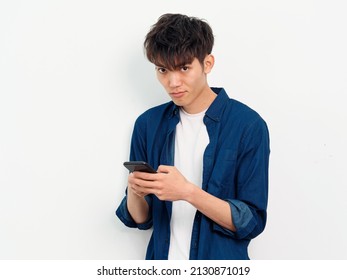 Portrait Of Handsome Chinese Young Man With Curly Black Hair In Blue Shirt Posing Against White Wall Background. Looking At Camera With Mobile Phone In Both Hands, Front View Studio Shot.