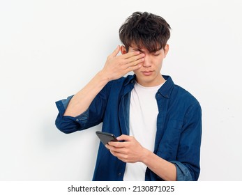 Portrait Of Handsome Chinese Young Man With Black Curly Hair In Blue Shirt Posing Against White Wall Background. Fingers Rubbing Eyes With Mobile Phone In Hand, Looks Tired, Front View Studio Shot.