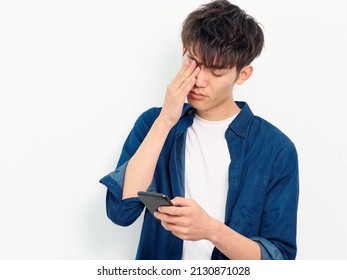 Portrait Of Handsome Chinese Young Man With Black Curly Hair In Blue Shirt Posing Against White Wall Background. Fingers Rubbing Eyes With Mobile Phone In Hand, Looks Tired, Front View Studio Shot.