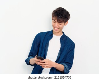 Portrait Of Handsome Chinese Young Man With Curly Black Hair In Blue Shirt Posing Against White Wall Background. Smiling And Looking At His Smartphone Holding In Both Hands, Front View Studio Shot.