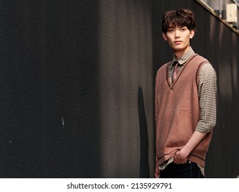 Portrait Of Handsome Chinese Young Man With Curly Black Hair In Plaid Shirt And Wool Vest Standing Against Black Wall Background In Sunny Day, Male Fashion, Cool Asian Young Man Lifestyle.