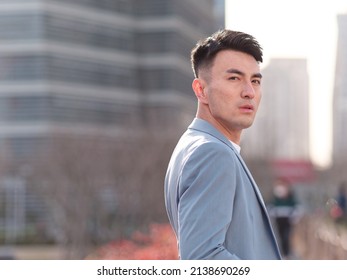 Portrait Of Handsome Chinese Young Man In Light Blue Suit Looking At Camera With City Buildings Background, Side View Of Confident Businessman.