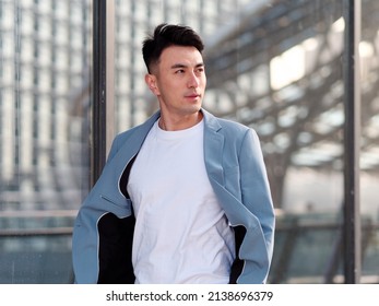 Portrait Of Handsome Chinese Young Man In Light Blue Suit And White Undershirt Walking And Looking Away With Modern City Buildings Background In Sunny Day, Front View Of Confident Businessman.