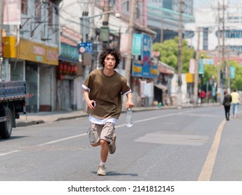 Portrait Of Handsome Chinese Young Man With Black Curly Hair In Brown T-shirt And Pants Running On Shanghai Old Town Street With Bottle Of Water In Hand, Front View Of Cool Young Man.