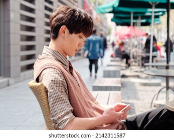 Portrait Of Handsome Chinese Young Man With Curly Black Hair In Plaid Shirt And Wool Vest Sitting Outdoor Cafe With His Mobile Phone In Hand In Sunny Day, Male Fashion, Cool Asian Young Man Lifestyle