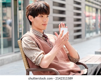Portrait of handsome Chinese young man with curly black hair in plaid shirt and wool vest sitting outdoor cafe with his mobile phone in hand in sunny day, male fashion, cool Asian young man lifestyle