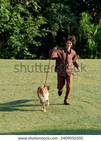 Portrait of handsome Chinese young man with curly hair running with his rough collie dog on green grass field in sunny day, male fashion, cool Asian young man lifestyle, harmony man and pet.