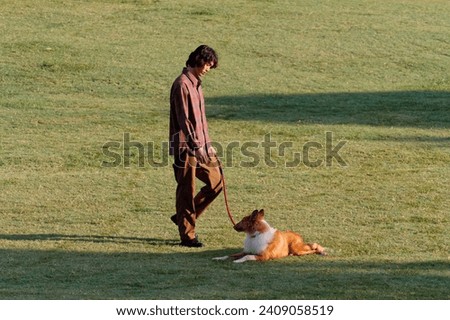 Portrait of handsome Chinese young man with curly hair walking with his rough collie dog on green grass field in sunny day, male fashion, cool Asian young man lifestyle, harmony man and pet.
