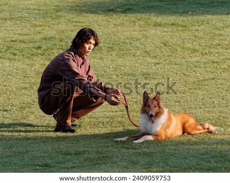 Portrait of handsome Chinese young man with curly hair squat with his rough collie dog on green grass field in sunny day, male fashion, cool Asian young man lifestyle, harmony man and pet.