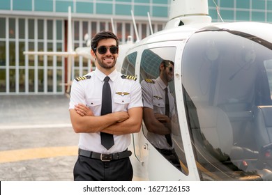 Portrait of handsome commercial pilot in captain white uniform standing and looking smart near small private helicopter on a landing point with private jet terminal at international airport