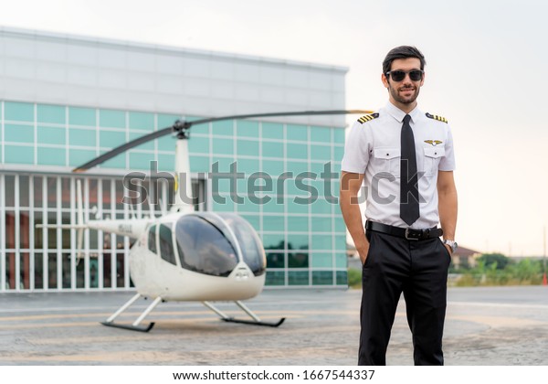 Portrait\
of handsome commercial pilot in captain white uniform standing and\
looking smart near small private helicopter on a landing point with\
private jet terminal at international\
airport.\
