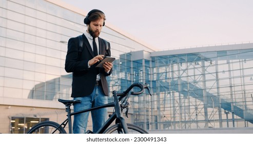 Portrait of handsome concentrated young bearded businessman in earphones which standing near his bike on the modern urban building background and using tablet pc - Powered by Shutterstock