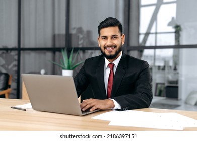 Portrait Of Handsome Confident Arabian Or Indian Successful Businessman, Entrepreneur, Lawyer, In A Formal Suit, Sit At Work Desk With Laptop In Modern Creative Office, Looks At Camera, Smile