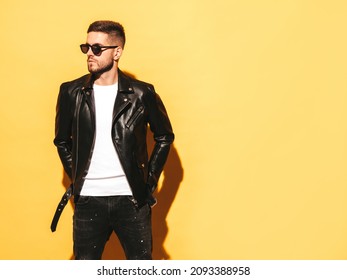 Portrait Of Handsome Confident Model. Sexy Stylish Man Dressed In Biker Leather Jacket And Black Jeans. Fashion Hipster Male Posing Near Yellow Wall In Studio In Sunglasses