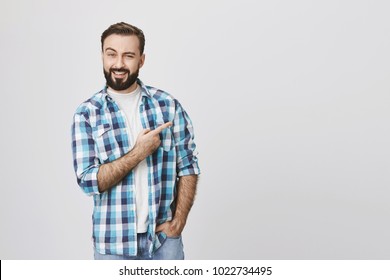 Portrait Of Handsome Confident Person With Beard, Lifting One Eyebrow And Smiling While Pointing Right, Wearing Casual Clothes And Standing Against Gray Background. Man Offers To Check Out New Bar