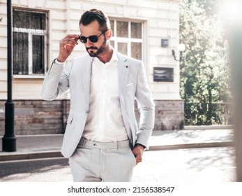 Portrait of handsome confident stylish hipster lambersexual model.Sexy modern man dressed in elegant white suit. Fashion male posing in the street background in Europe city at sunset. In sunglasses