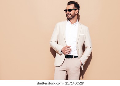 Portrait Of Handsome Confident Stylish Hipster Lambersexual Model. Sexy Modern Man Dressed In White Elegant Suit. Fashion Male Posing In Studio Near Beige Wall In Sunglasses