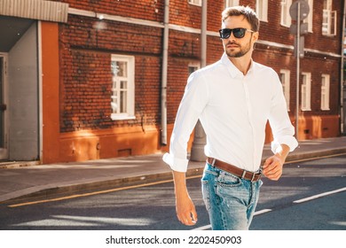 Portrait of handsome confident stylish hipster lambersexual model.Modern man dressed in white shirt. Fashion male posing on the street background in sunglasses. Outdoors at sunset. Walking