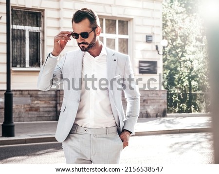 Portrait of handsome confident stylish hipster lambersexual model.Sexy modern man dressed in elegant white suit. Fashion male posing in the street background in Europe city at sunset. In sunglasses
