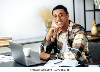 Portrait Handsome Confident Successful Young Mixed Race Latino Man, Freelancer Or Manager, Wearing Shirt And Glasses, Sits At Work Desk With Laptop, Looking At The Camera And Smiling Friendly