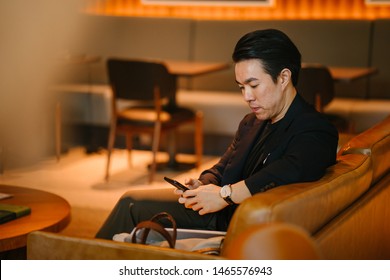 Portrait of a handsome, confident and well-dressed young Chinese Asian man checking his smartphone as he sits on a leather sofa in a well-appointed sofa during the day. 