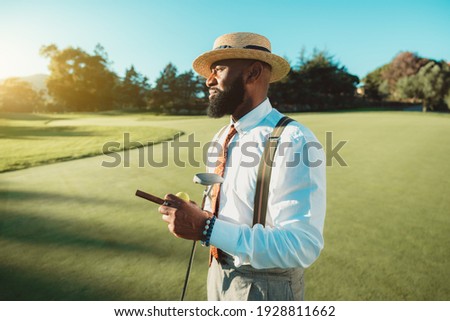 A portrait of a handsome dapper bearded black man in an elegant outfit: white shirt, trousers with suspenders, tie, and hat, is standing on the golf field and holding the club, cigar, and ball in hand