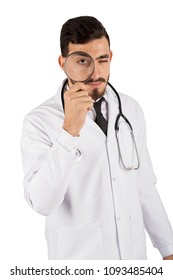 Portrait Of A Handsome Doctor Wearing His Uniform, Standing And Holding A Magnifying Glasses Searching For Something, Isolated On White Background