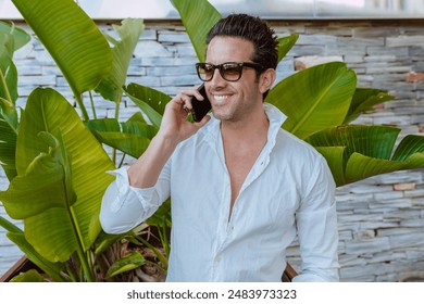 Portrait of a handsome elegant businessman wears white shirt and sunglasses, talking on smartphone, broadly smiles looking away, posing in front of a plan with big leafs and stone wall. - Powered by Shutterstock