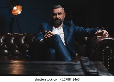 Portrait of handsome elegant mature man with cigar and a glass whiskey in a dark room