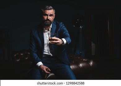 Portrait Of Handsome Elegant Mature Man With Cigar And A Glass Whiskey In A Dark Room