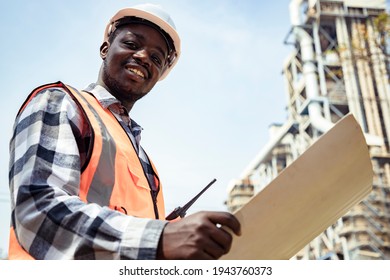 Portrait Of Handsome Engineering Man Holding Paperwork And Walkie Talkie With Wear Hardhat In Front Of Oil Industry Factory. Back View Of Contractor On Background Of Modern Buildings.