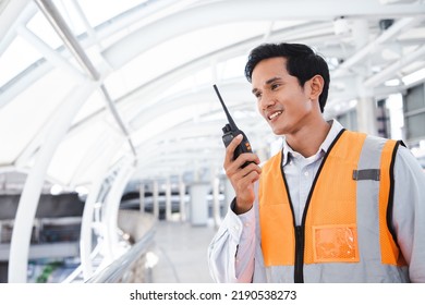 Portrait Of Handsome Engineering Man Using Walkie Talkie And Wear Hardhat In Front Of Oil Industry Factory. Back View Of Contractor On Background Of Modern Buildings.