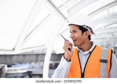 Portrait of handsome engineering man using walkie talkie and wear hardhat in front of construction outside work station. Back view of contractor on background of modern buildings.