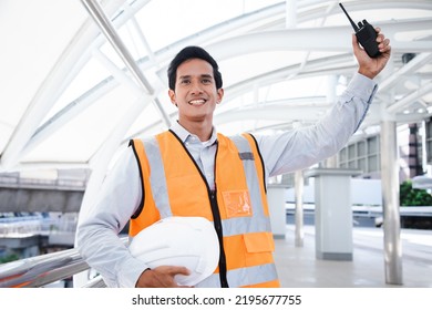 Portrait Of Handsome Engineering Man Using Walkie Talkie And Wear Hardhat In Front Of Oil Industry Factory. Back View Of Contractor On Background Of Modern Buildings.