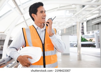 Portrait Of Handsome Engineering Man Using Walkie Talkie And Wear Hardhat In Front Of Oil Industry Factory. Back View Of Contractor On Background Of Modern Buildings.