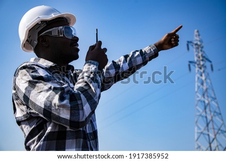 Portrait of handsome engineering man using walkie talkie and wear hardhat in front of High power power plant. Back view of contractor on background of modern buildings.