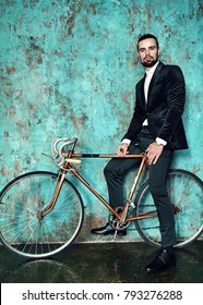 portrait of handsome fashion stylish hipster lumbersexual businessman model dressed in elegant black suit posing near sport bicycle and blue grange wall in studio. Metrosexual