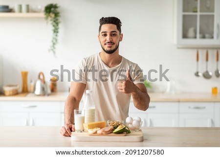 Portrait of handsome fit young Arab man offering healthy products rich in protein and showing thumb up gesture at kitchen. Middle Eastern guy recommending wholesome nutrition at home