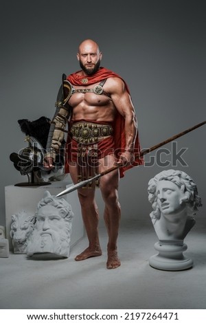 Portrait of handsome gladiator with spear posing around marble greek statues.