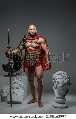 Portrait of handsome gladiator with spear posing around marble greek statues.