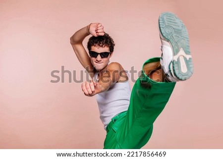 Portrait of handsome guy, throwing high kick in the air, practicing self defense kicking confident facial expression wear sun specs, white t-shirt and green pants isolated over beige color background.