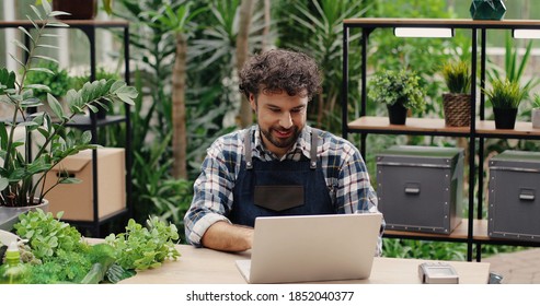 Portrait Of Handsome Happy Caucasian Male Professional In Apron Sitting At Desk In Floral Store And Typing On Laptop. Joyful Young Man Florist Working On Computer At Workplace. Small Business Concept